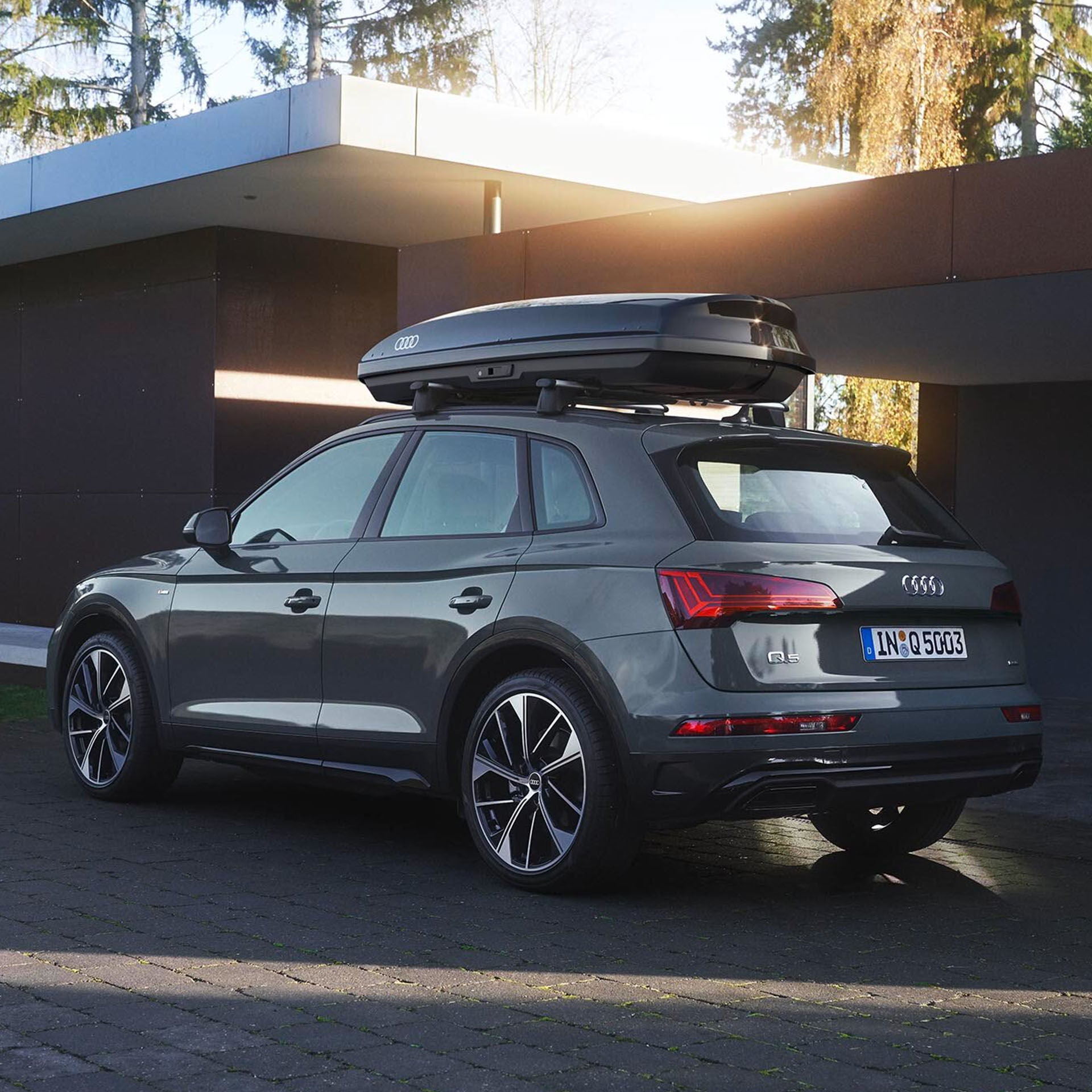 Ein grauer Audi Q5 mit Dachbox steht vor einem modernen Gebäude, Sonnenlicht fällt durch Bäume im Hintergrund.