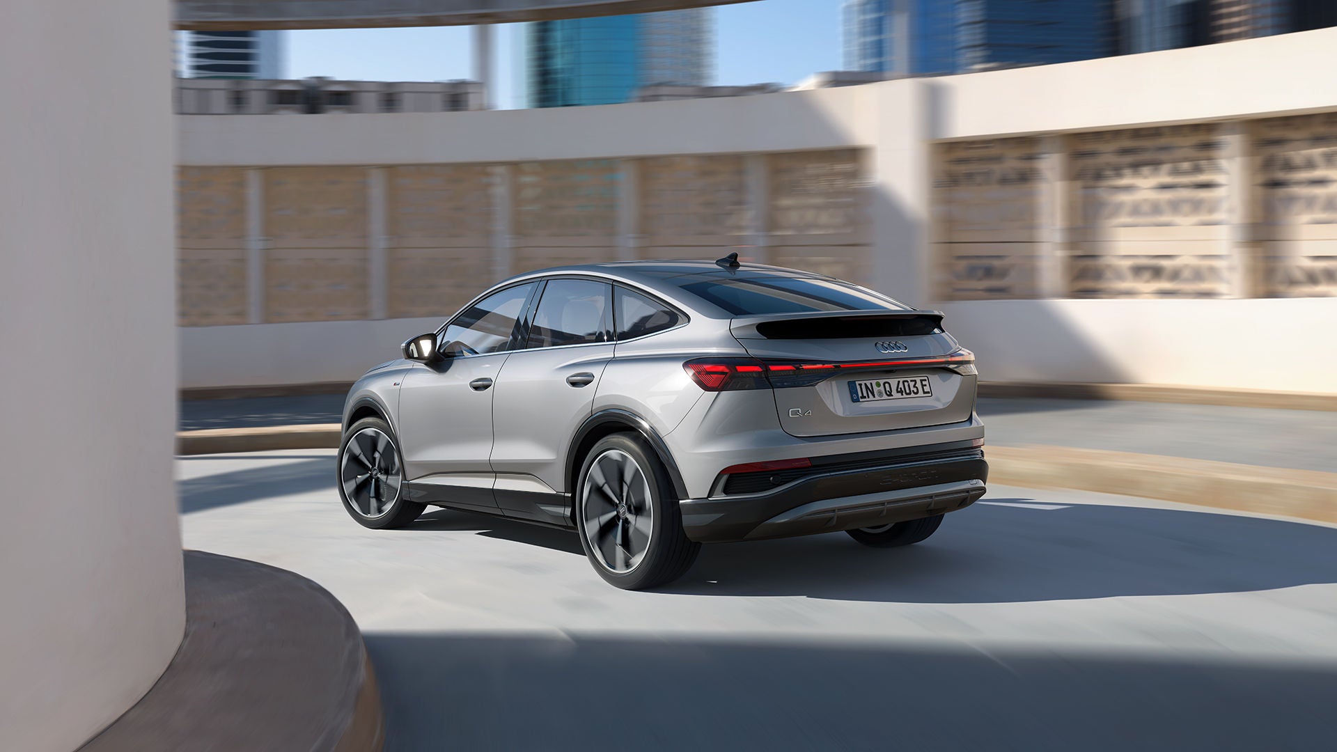 Silberner Audi Q4 e-tron fährt auf einer Straße mit moderner Architektur im Hintergrund, Seiten- und Heckansicht des Fahrzeugs.