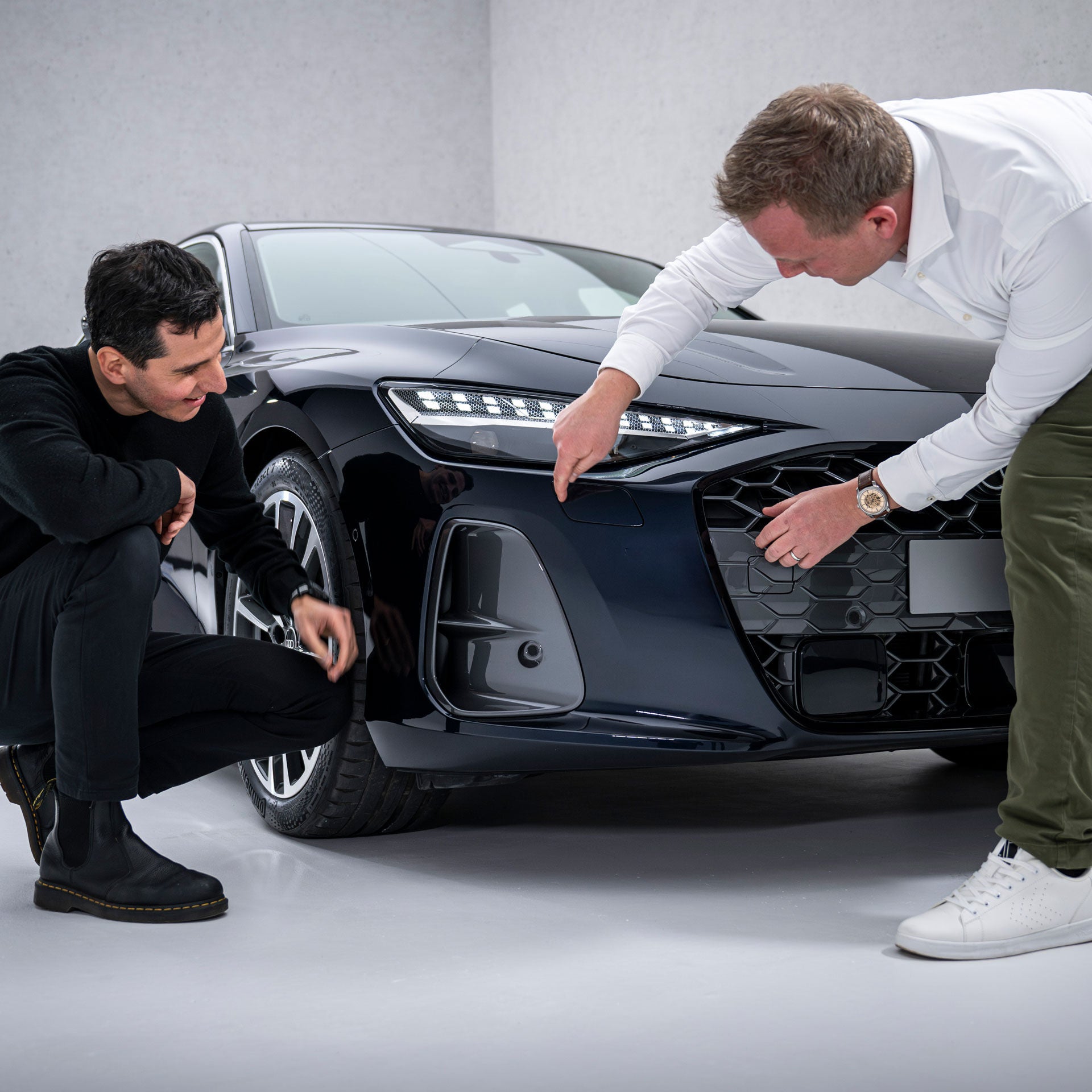 Francesco d’Amore (kniend) und Sebastian Weiper (gebückt) stehen vor der Front einer schwarzen Audi A6 Limousine und sehen sich die seitlichen Lufteinlässe, die sogenannten Air Curtains, an.