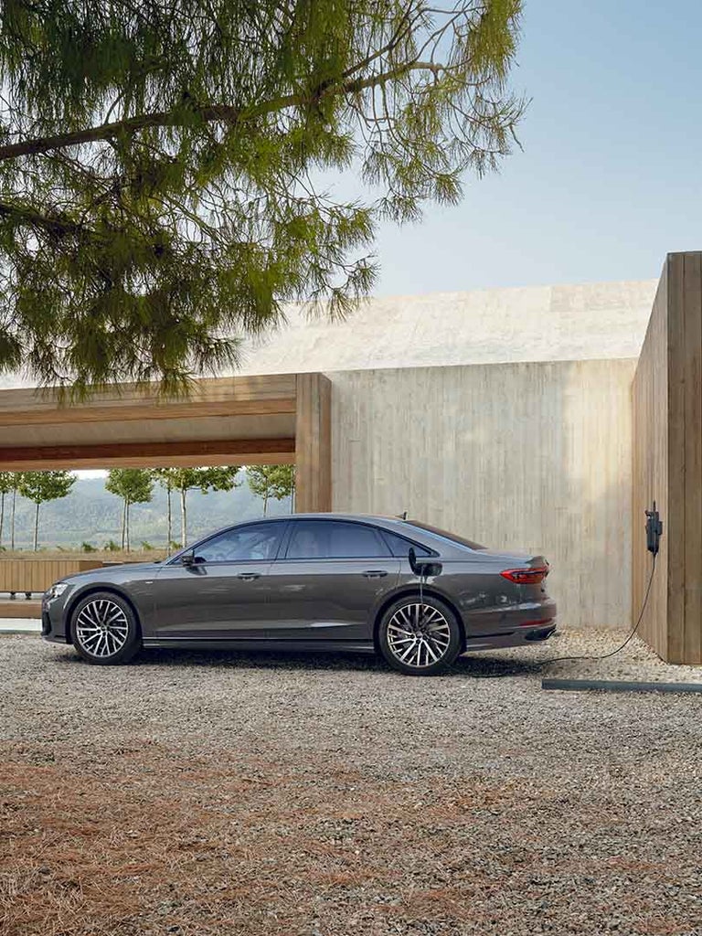 Ein grauer Audi A8 L 60 TFSI e lädt an einer Wandladestation in naturnaher Umgebung mit hellem Holzgebäude. Weit im Hintergrund steht ein farbiger Mann.