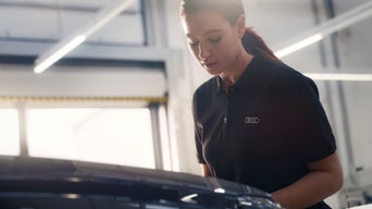 Frau im Audi-Poloshirt arbeitet in einer Werkstatt an einem Fahrzeug, helle Halle im Hintergrund.