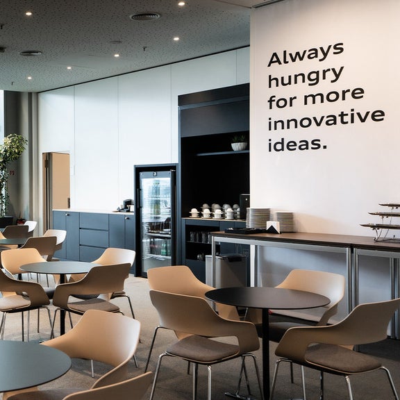 Cafeteria mit runden Tischen und Stühlen, Glasfront und Schriftzug an der Wand.