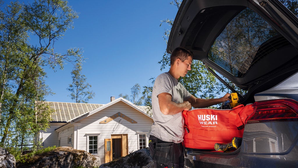 André Myhrer tar ut en väska från hans klädmärke "Huski wear" ur backluckan av sin bil, i bakgrunden syns huset han bygger