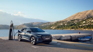 The Q4 SUV e-tron, viewed from the front at an angle, speeding down a country road alongside a forest lake, with a mountainous landscape in the background.
