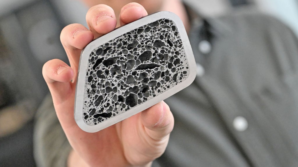 Hand hält eine rechteckige Materialprobe mit poröser schwarz-grauer Struktur.