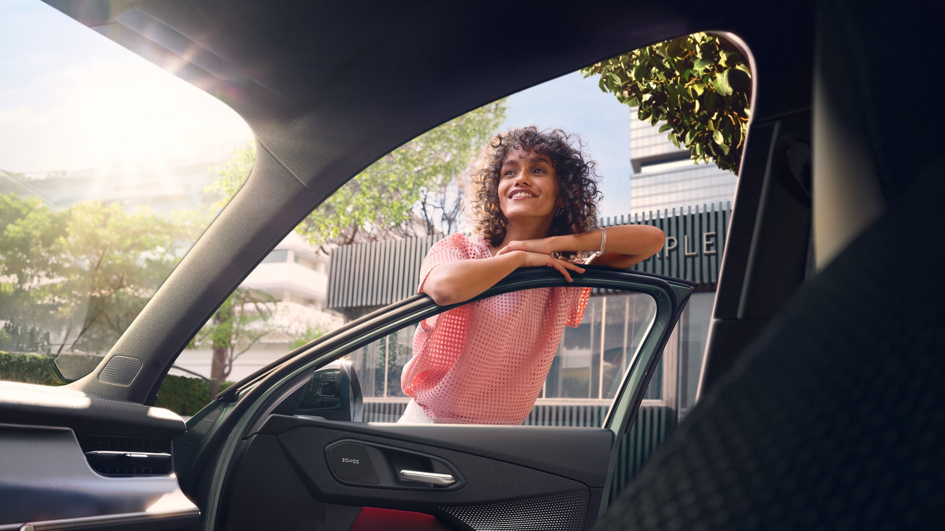 Eine Frau mit gelockten Haaren lehnt an einer offenen Autotür und wird aus dem Innenraum heraus aufgenommen.