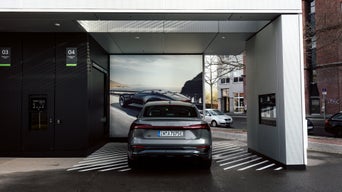 Audi-Elektroauto lädt an urbaner Audi-Ladestation mit großem Plakat und markierten Ladeflächen bei Tageslicht.
