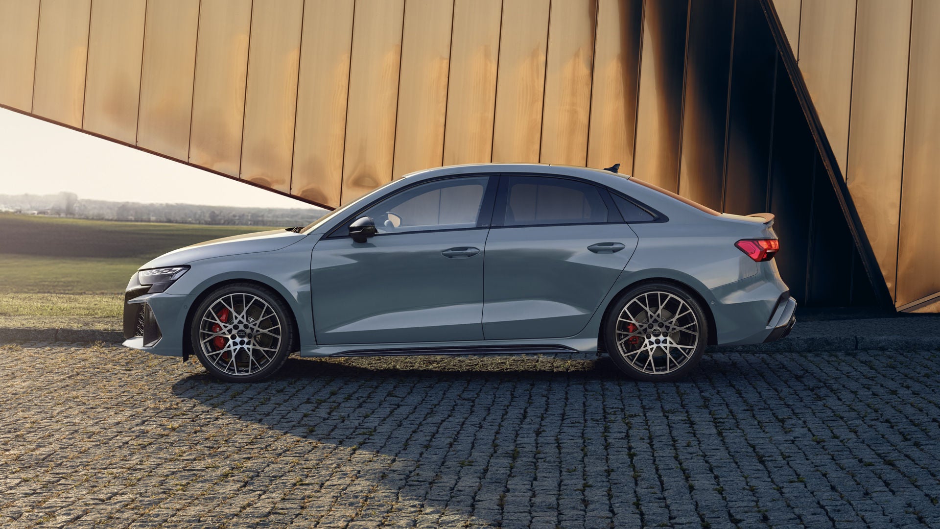 Die RS 3 Limousine von der Seite parkend auf Kopfsteinpflaster, im Hintergrund Umrisse eines modernen Gebäudes.