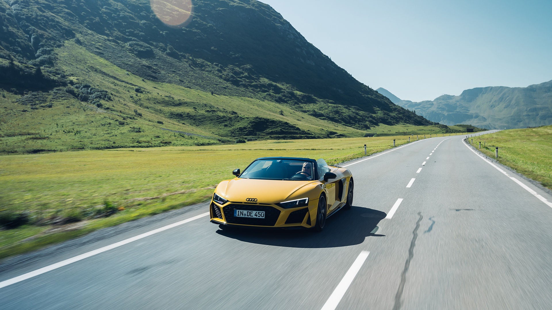 A yellow Audi convertible drives along a country road through green mountain scenery under a blue sky.