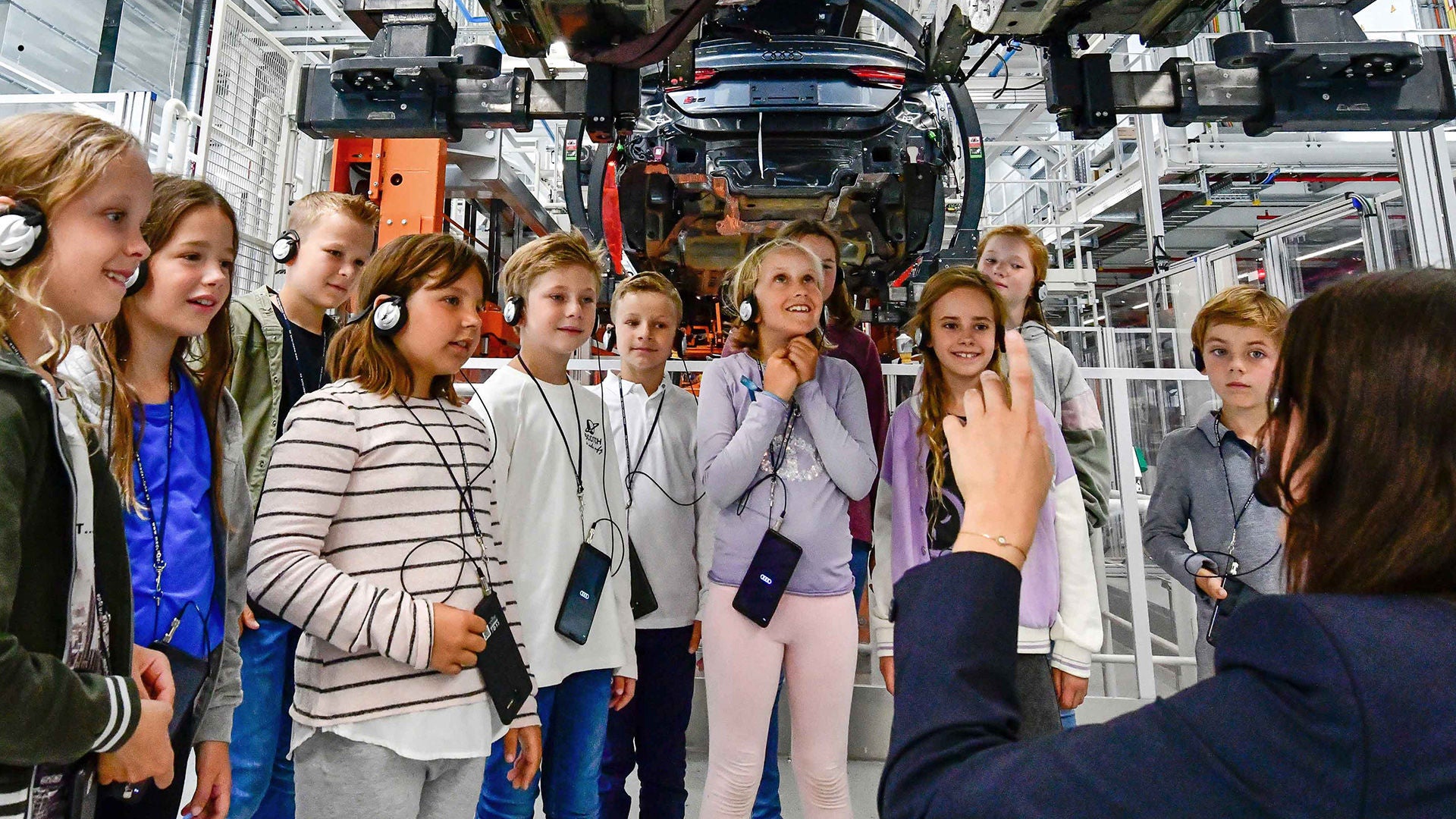 Children with headphones on a tour of the plant, looking excitedly at vehicle assembly above their heads.