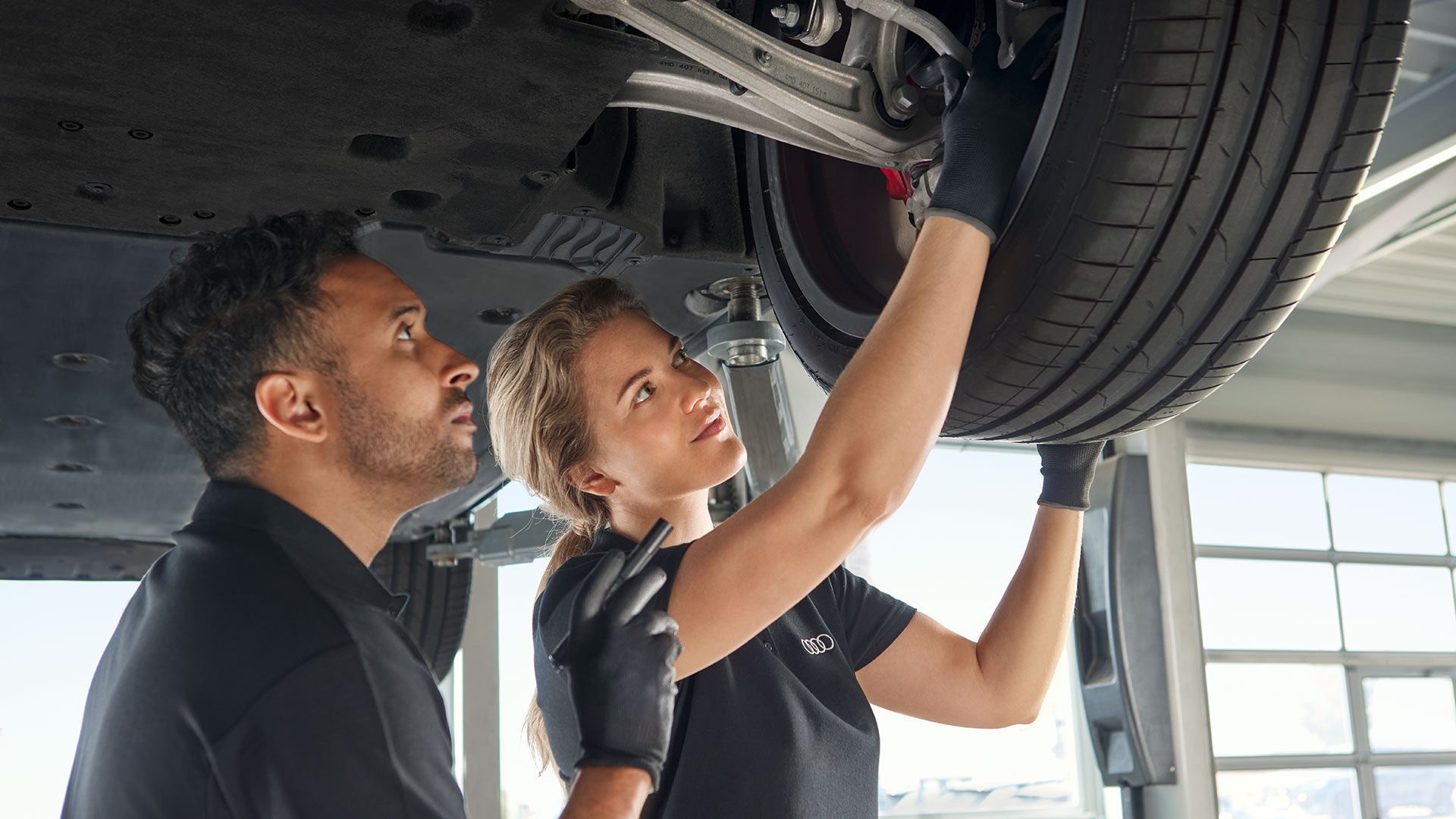 Zwei Audi-Mitarbeitende prüfen gemeinsam ein angehobenes Fahrzeugrad in einer Werkstatt, beide tragen schwarze Handschuhe.