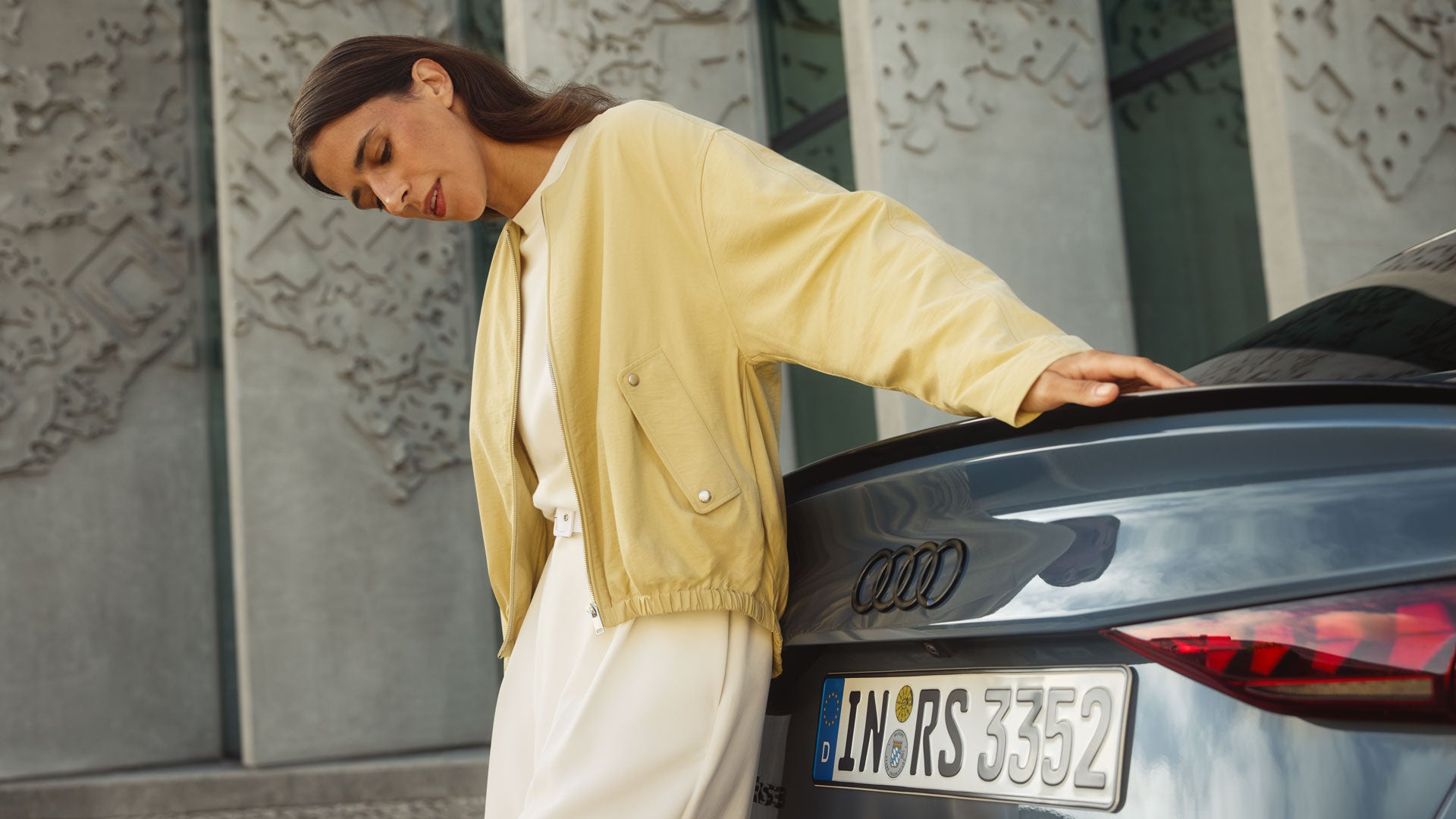 Frau lehnt sich in heller Kleidung entspannt an das Heck einer Audi RS3 Limousine, im Hintergrund ein modernes Gebäude.