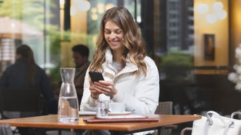 Eine junge Frau sitzt an einem Tisch in einem Café, blickt lächelnd auf ihr Smartphone.