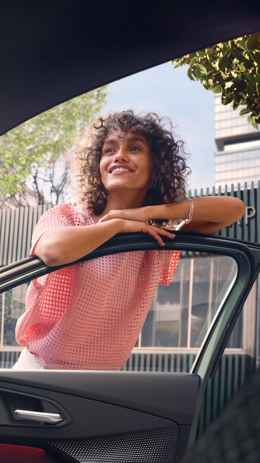 Eine Frau mit gelockten Haaren lehnt an einer offenen Autotür und wird aus dem Innenraum heraus aufgenommen.