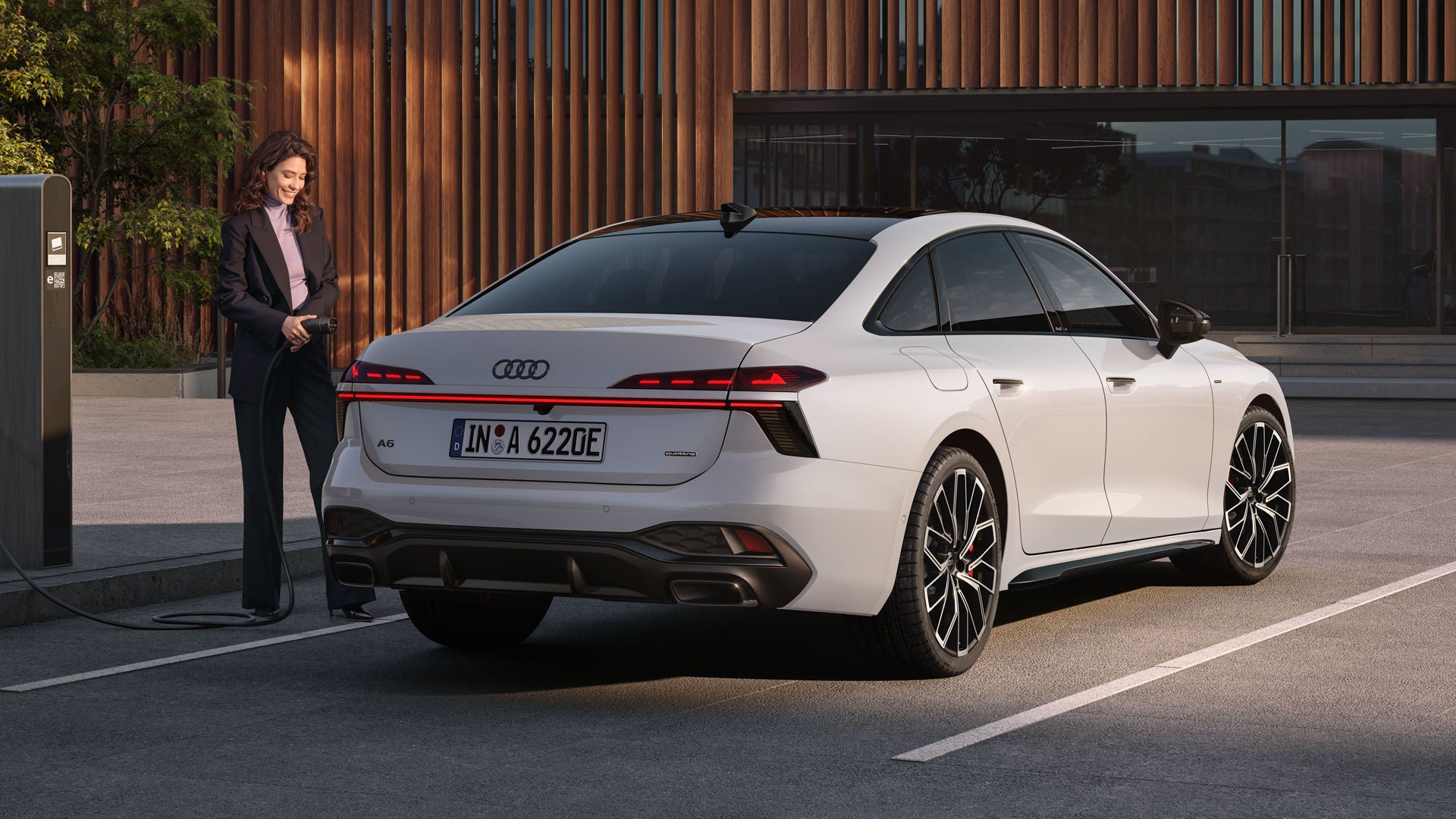 Die A6 Limousine e-hybrid in weiß steht auf einem Parkplatz vor einer Gebäudefassade. Eine junge Frau schließt die A6 Limousine e-hybrid an eine Ladestation an.