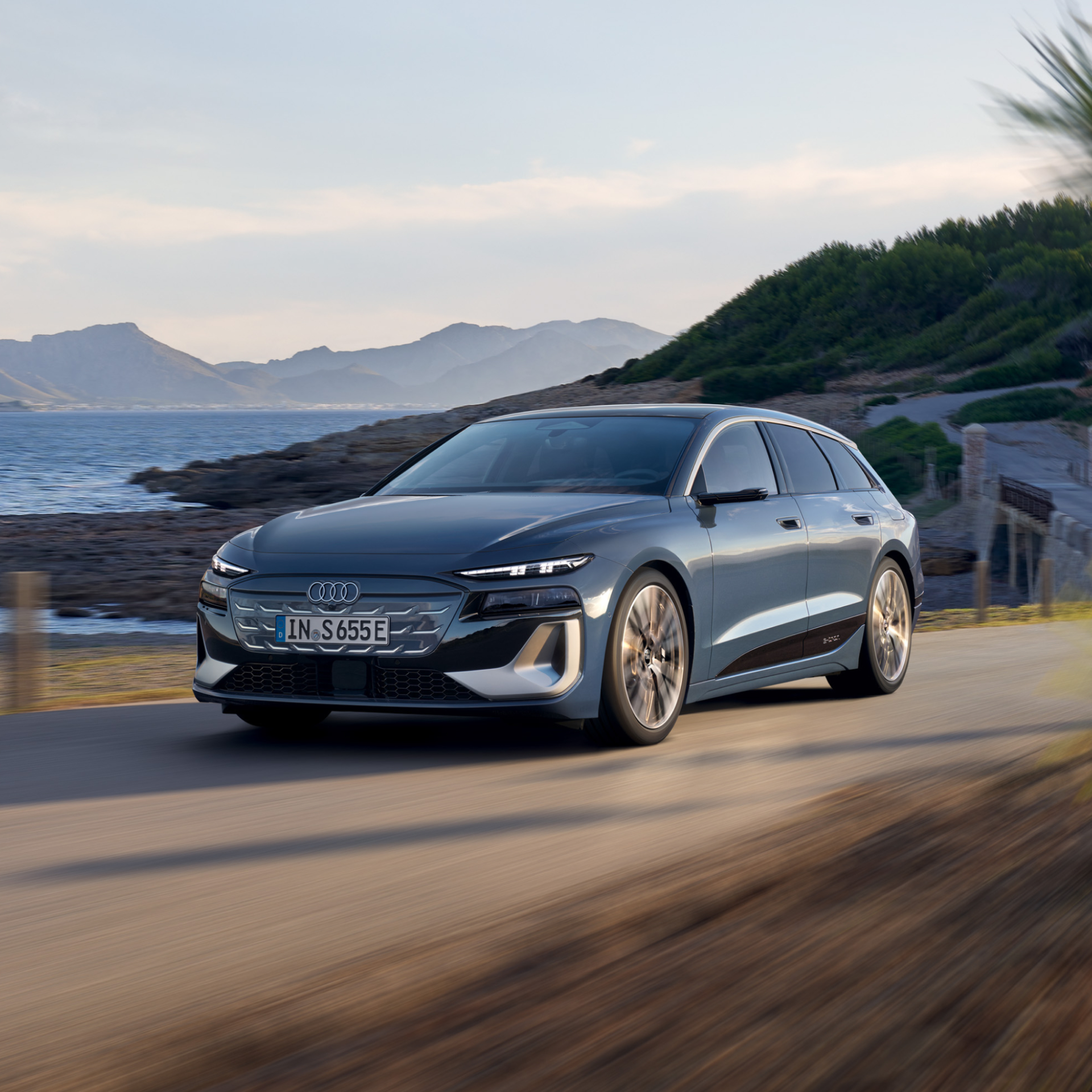 A blue Audi S6 Avant e-tron cruising on a coastar road with water and mountains in the background