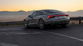 A silver Audi RS e-tron GT drives along a winding road at sunset, with glowing taillights and a mountain backdrop.