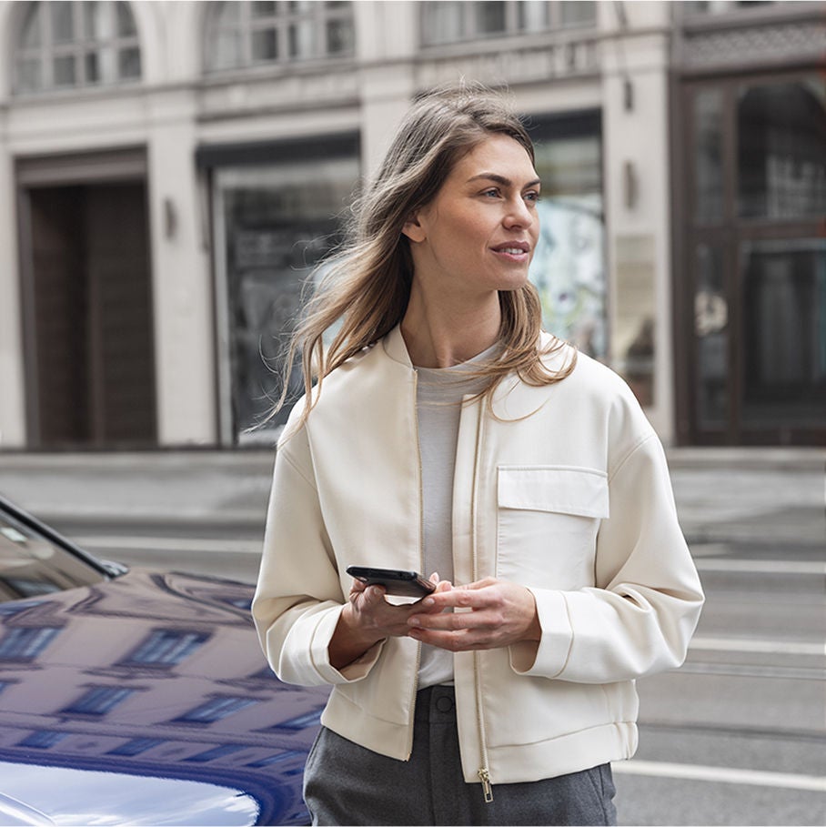 A woman in a cream bomber jacket stands on a street, holding a smartphone, with an urban backdrop of buildings and a car.