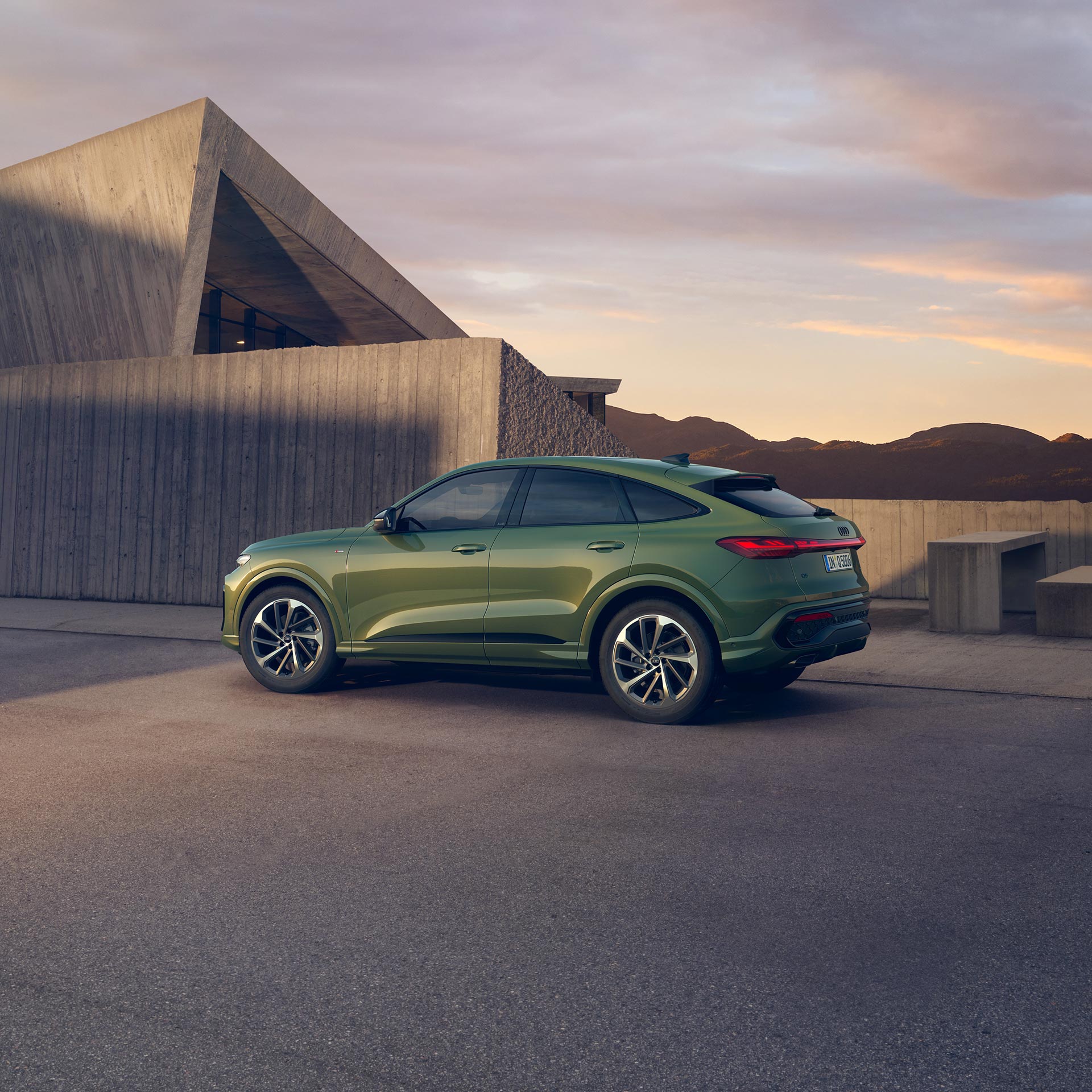 Ein grüner Audi Q5 Sportback steht vor modernem Betongebäude bei tief stehender Sonne. Hinter dem Gebäude sind Berge zu sehen.