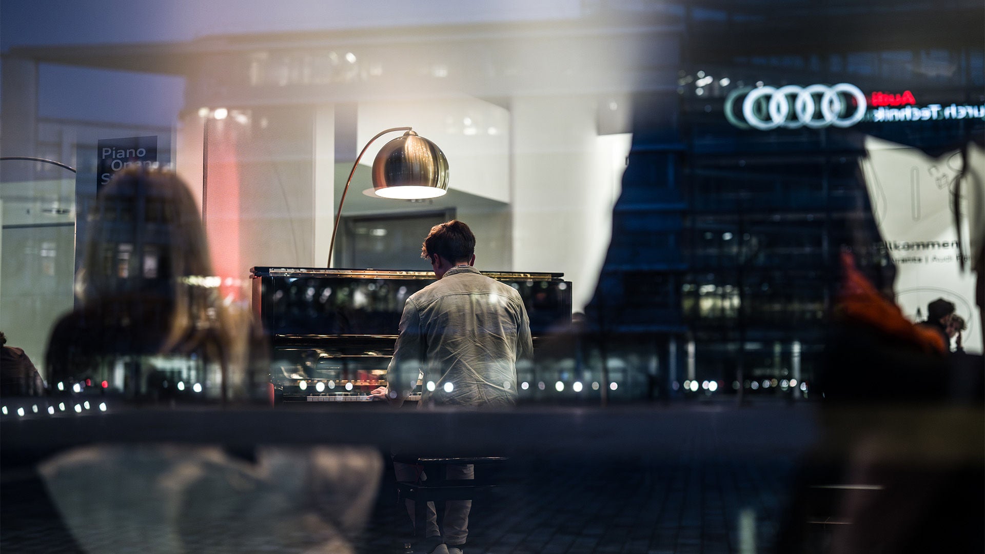 Mann spielt Klavier in beleuchtetem Innenraum, durch Fenster mit Spiegelungen und Audi-Logo im Hintergrund.