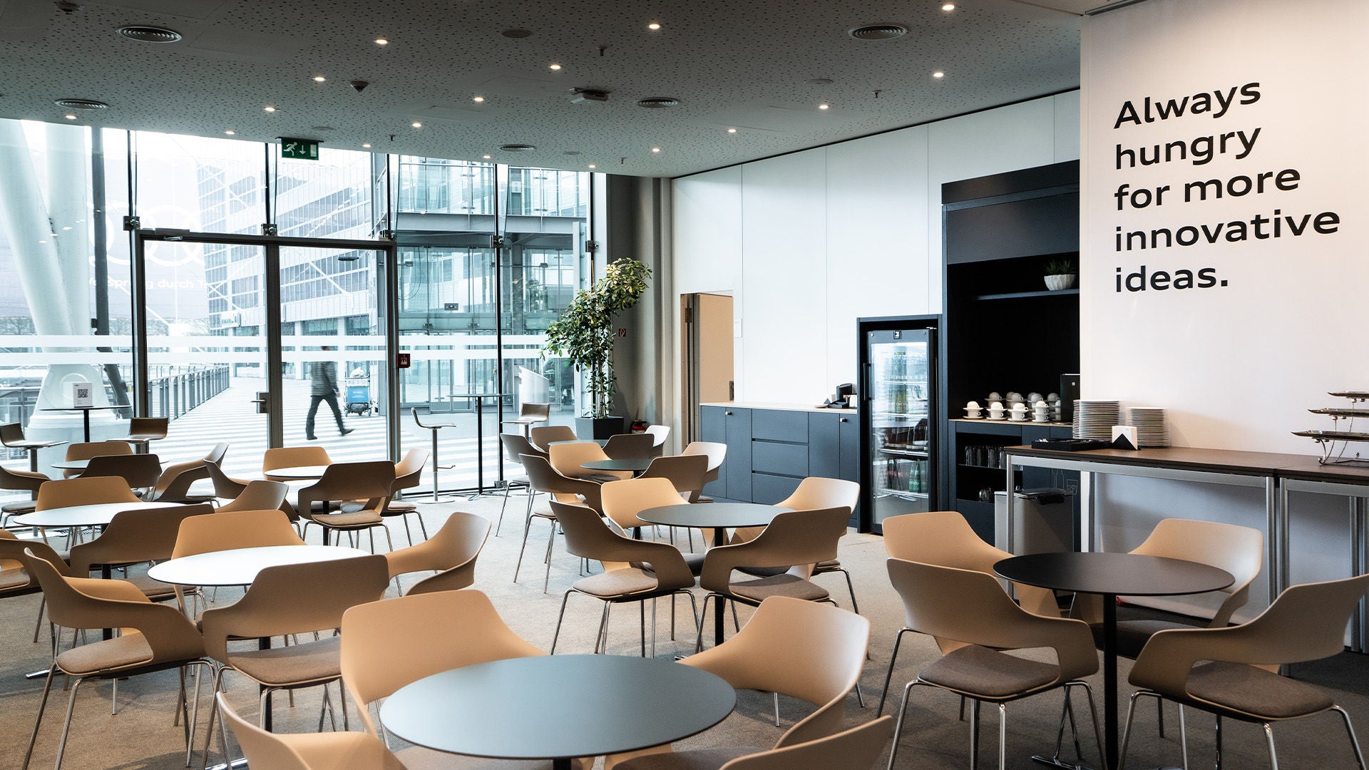 Heller Aufenthaltsbereich mit Tischen, Holzstühlen, Kaffeestation und Wandtext „Always hungry for more innovative ideas.“