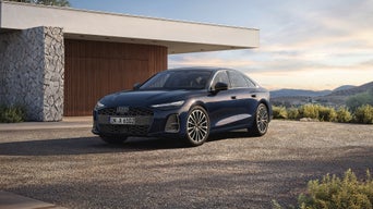 A blue Audi A6 Saloon parked outside a modern home, surrounded by gravel, with mountains and a sunset in the background.