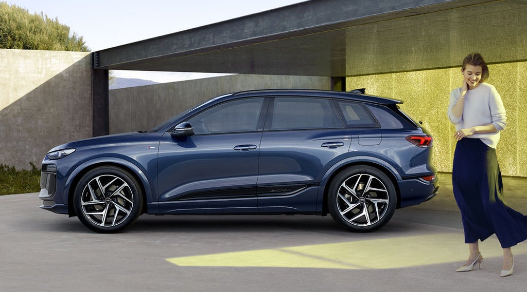 Den mörkblå Q6 SUV e-tron visas i profil framför en stadsbyggnad. I förgrunden, något åt sidan om bilen står en kvinna.