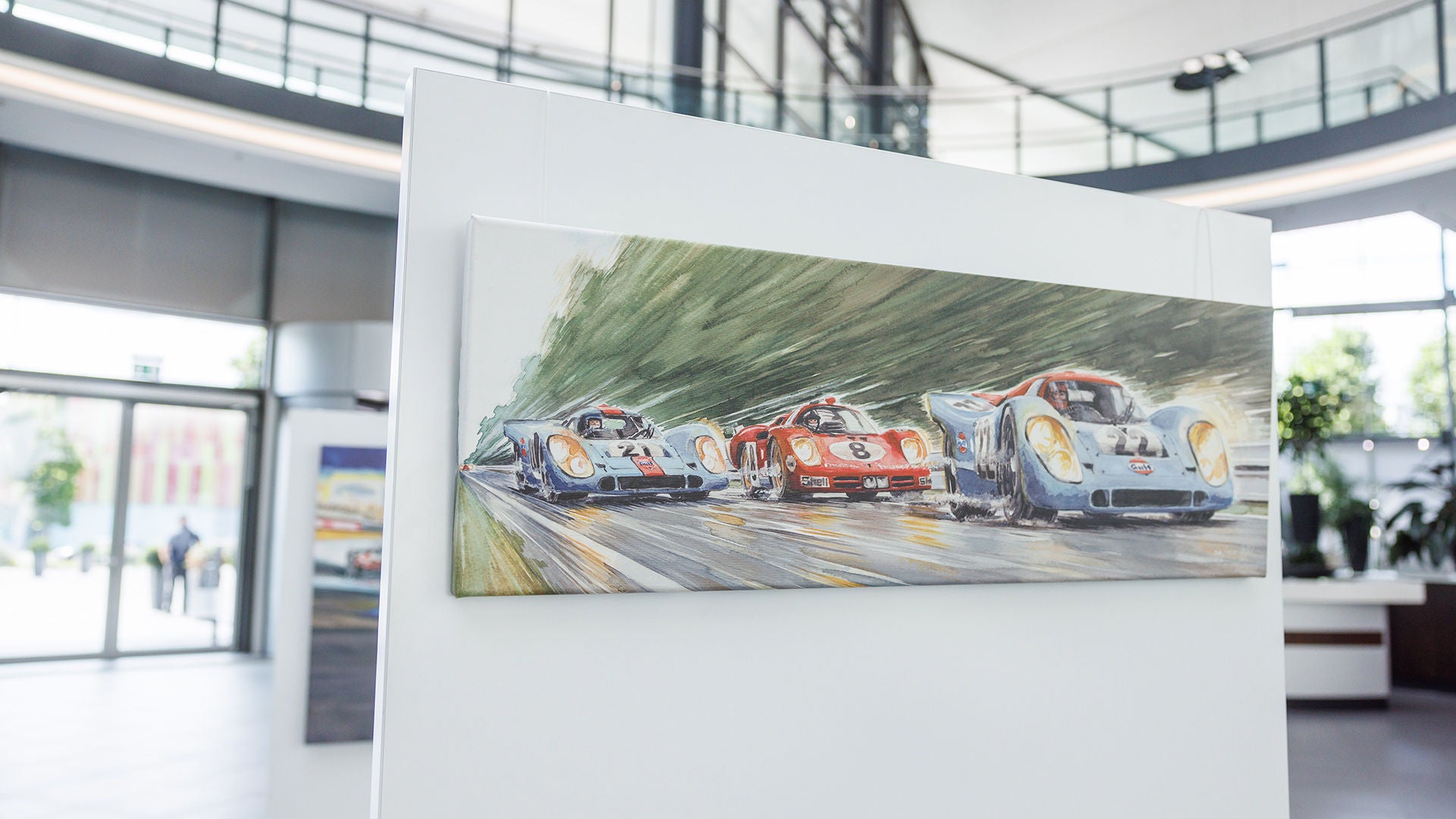 Aquarell-Gemälde mit drei Rennwagen auf einer Rennstrecke, ausgestellt auf einem weißen Ständer in einer hellen Empfangshalle.