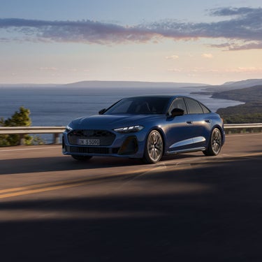 Blaue Audi S5 Limousine fährt auf Küstenstraße mit Meer und Bergen im Hintergrund.