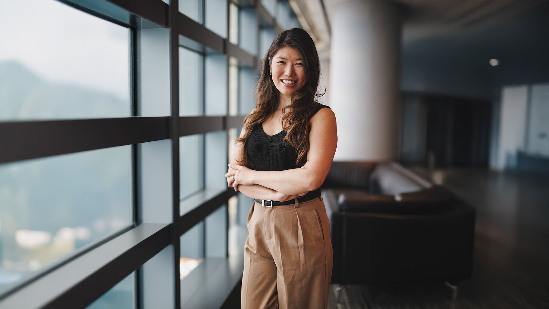 Frau mit langen Haaren im schwarzen Oberteil und beiger Hose, lächelnd, vor Fensterfront in modernem Gebäude.