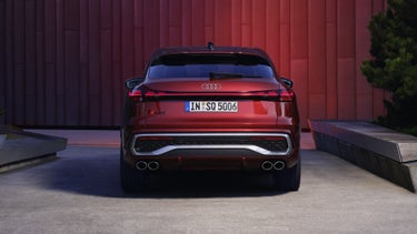 Heckprofil des roten SQ5 Sportback, welcher in einem Stadtpark vor einer Gebäudefassade steht und diese rot anleuchtet.