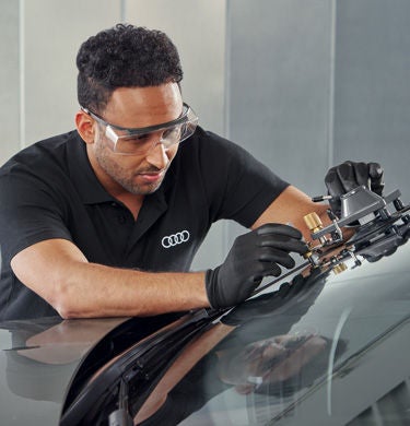 Mechaniker mit Schutzbrille repariert Windschutzscheibe eines Audi mit Spezialwerkzeug in der Werkstatt.