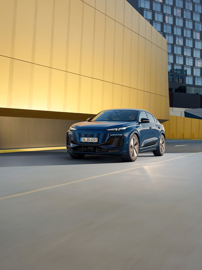 SQ6 Sportback e-tron kör på en väg. En gul byggnad syns i bakgrunden.
