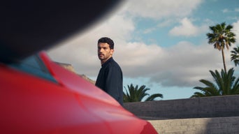 A man stands beside a red car, with palm trees and cloudy blue skies in the background, creating a serene outdoor setting.