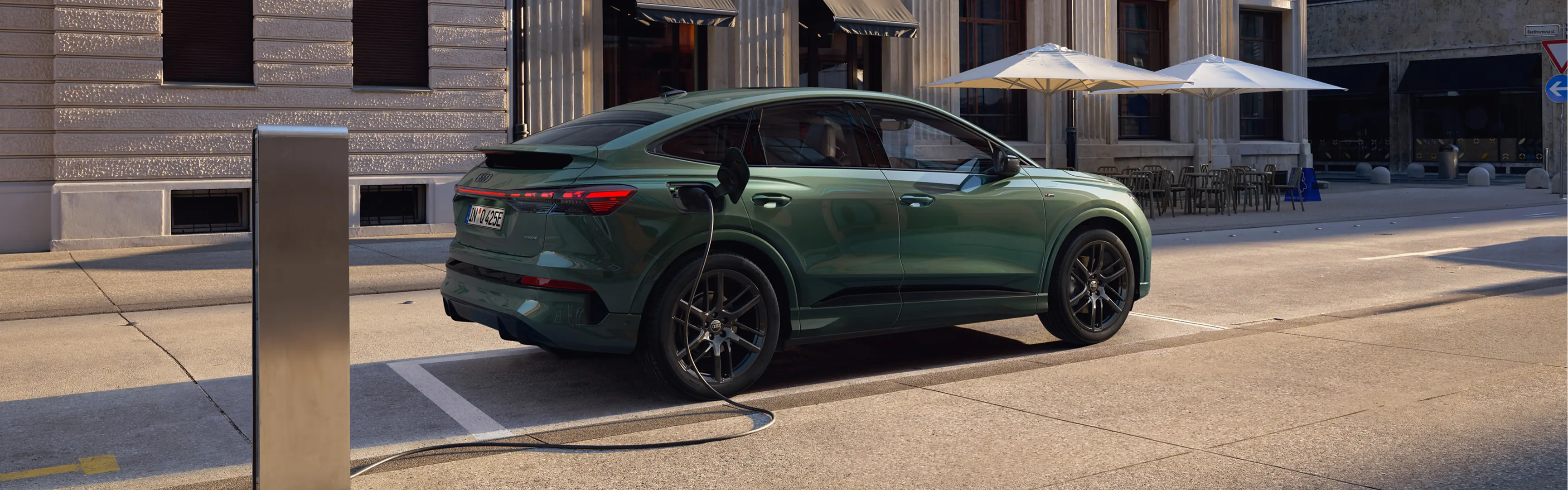 Green Audi Q6 Sportback e-tron connected to a public charging station on a city street, shown from the rear three-quarter angle. The vehicle features a sloping coupé-style roofline, large alloy wheels and a full-width rear LED light bar. Modern buildings, outdoor café seating and parasols form the urban backdrop, while daylight highlights the charging cable connected to the rear charging port.