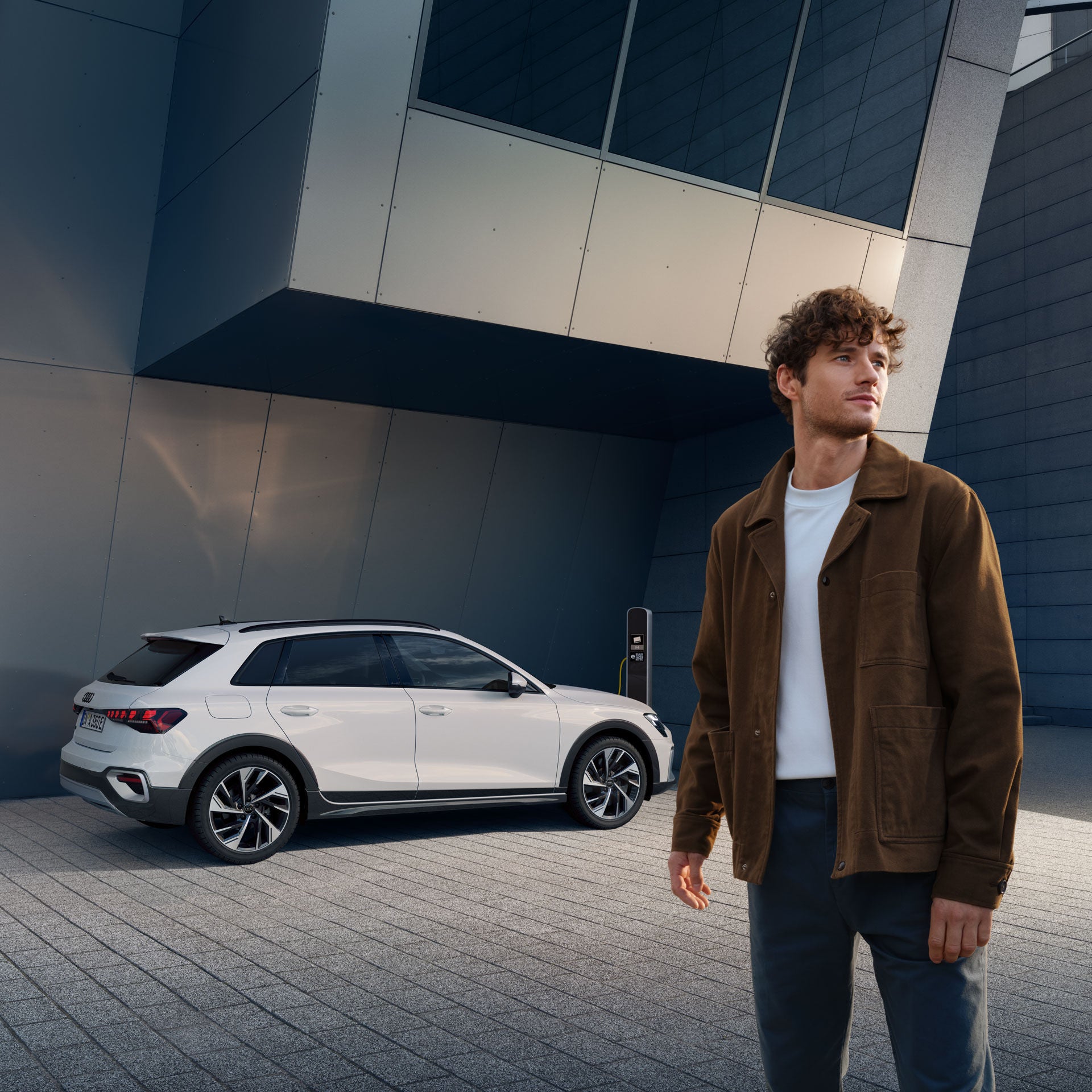 Mann steht vor einem weißen Audi A3 an einer Ladestation, moderner Gebäudekomplex mit Glas- und Metallfassade im Hintergrund.