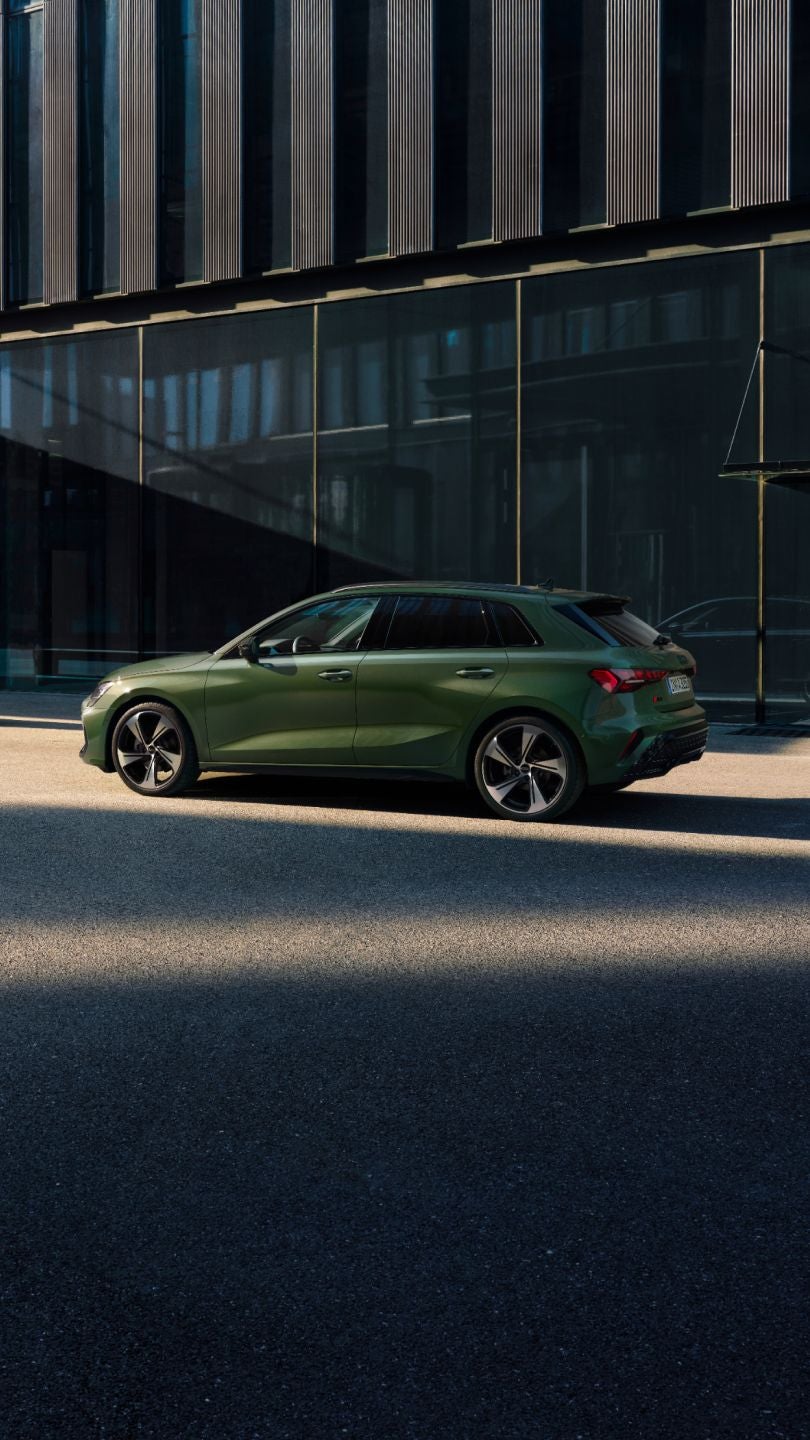 A green Audi A3 Sportback car parked beside glass buildings in sunlight.
