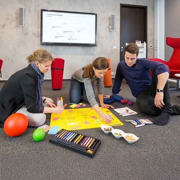 Drei Personen sitzen auf dem Boden in einem Büro, schreiben an Plakat, umgeben von Stiften und Snacks.