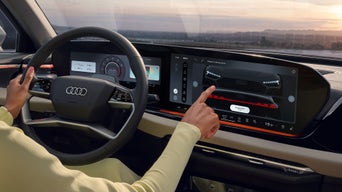 Fahrerin im gelben Outfit bedient großes Touchdisplay im Audi, Blick auf Digitalanzeigen und Landschaft bei Sonnenuntergang.