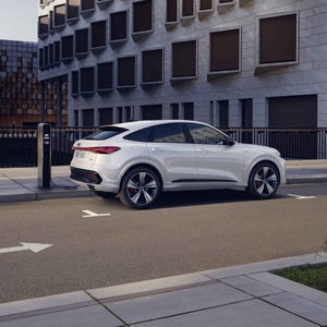 Vista laterale di Audi Q5 Sportback e‑hybrid bianca in fase di ricarica in un parcheggio davanti a un edificio alto.