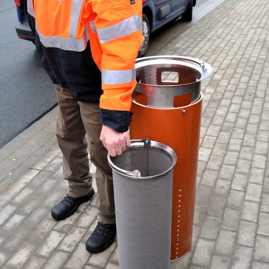 Man in a safety jacket stands on the sidewalk holding a cylindrical metal sieve next to an open filter system.