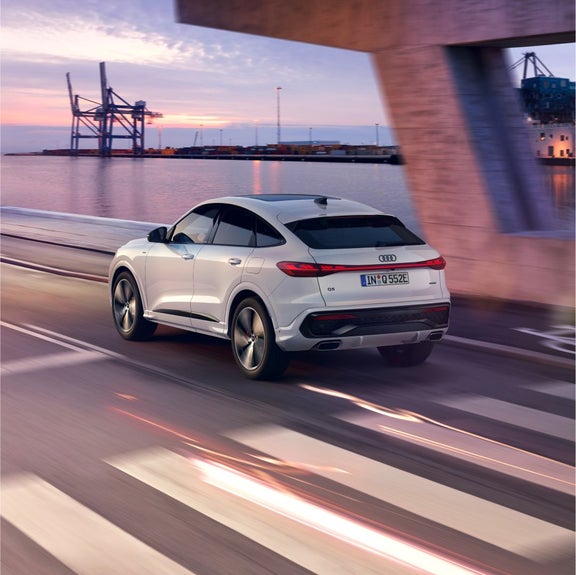 A white Audi Q5 drives along a waterfront road at sunset, with industrial cranes and shipping containers in the background.