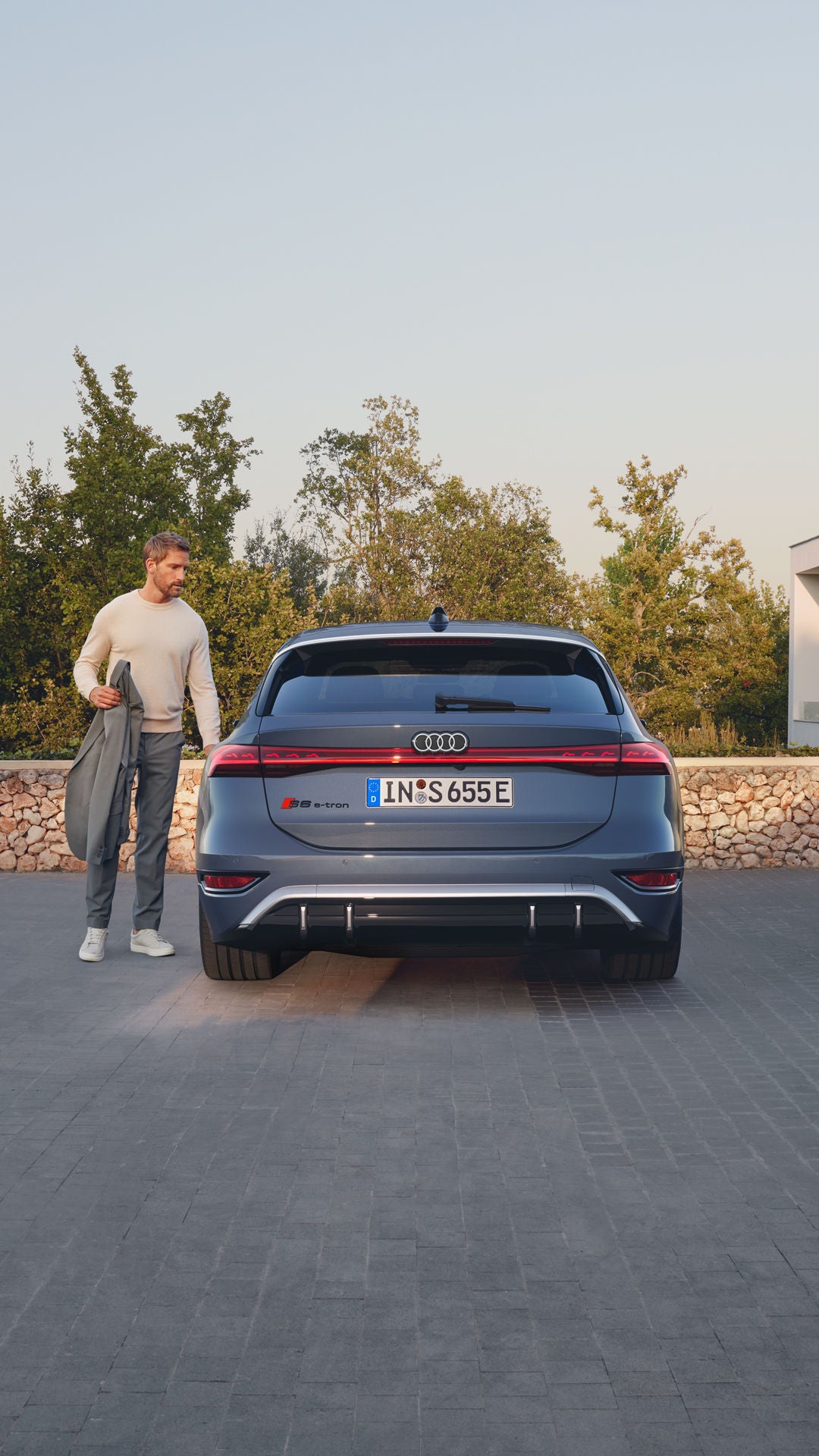 Heckprofil des S6 Avant e-tron, welcher auf Pflastersteinen vor einer Steinmauer und dem Garten eines modernen Hauses steht. Ein Mann mittleren Alters ist in Begriff, eine Tür zu öffnen.
