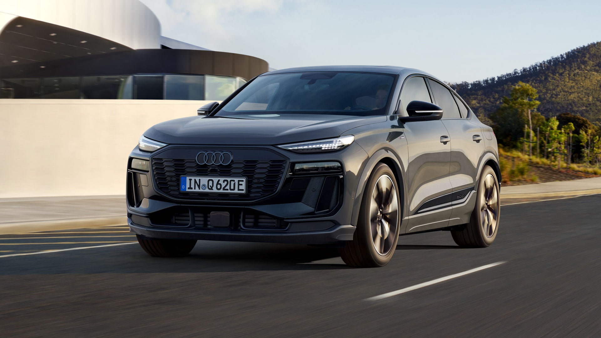 Schräges Frontprofil des Q6 Sportback e-tron, welcher auf einer Straße neben einem modernen Haus fährt.