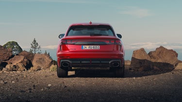 The RS Q8 SUV Performance is shown from behind, parked on a gravel road. Small rocks are visible on the left and right, with a few trees and a band of clouds in the background.