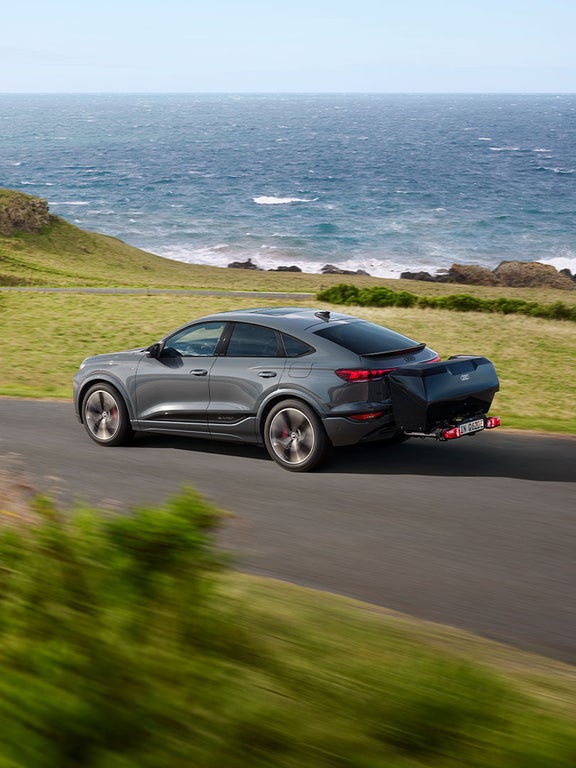 Grauer Audi Q6 Sportback e-tron mit Heckbox fährt auf Küstenstraße durch grüne Hügellandschaft am Meer.