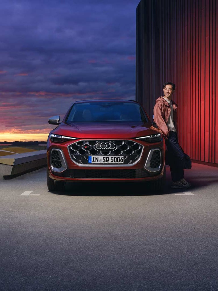 Ein roter Audi SQ5 Sportback frontal, daneben steht ein asiatischer Mann ans Auto gelehnt in Freizeitkleidung vor rot beleuchteter Wand, im Hintergrund geht die Sonne unter.