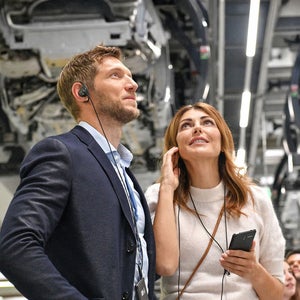 Mann und Frau mit Audioguide schauen in einer Fabrikhalle nach oben; die Frau hält ein Smartphone.