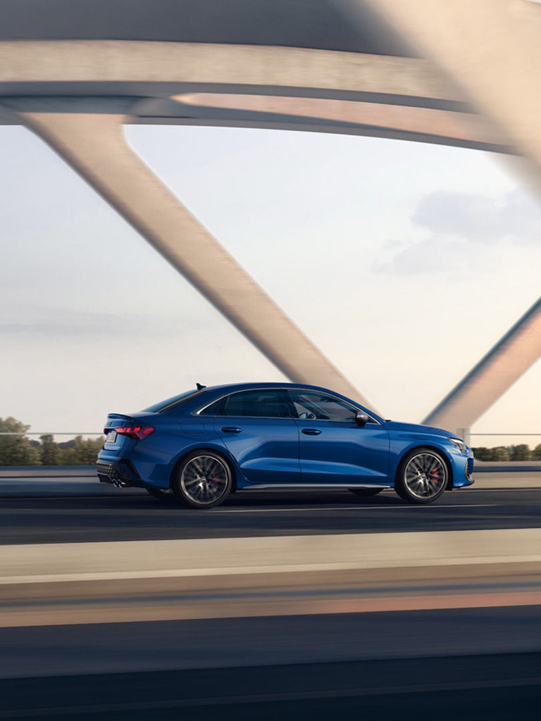Blaue Audi S3 Limousine fährt auf einer Brücke mit moderner Architektur bei Tageslicht.
