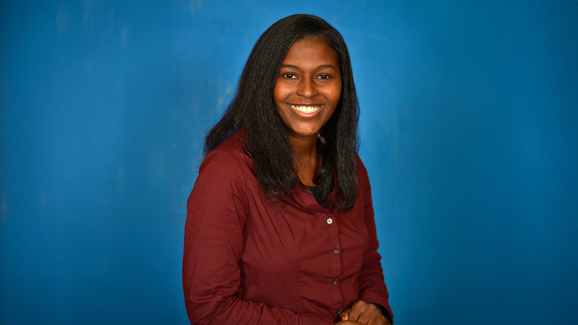 Frau mit langen schwarzen Haaren im bordeauxfarbenen Hemd, lächelnd, vor blauem Hintergrund.